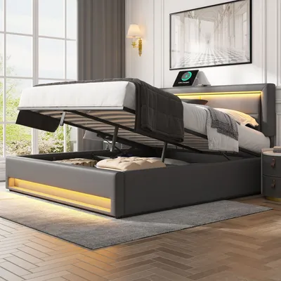 A modern bedroom with a grey upholstered bed frame with its storage compartment open, revealing stored items. The bed has integrated LED lighting.