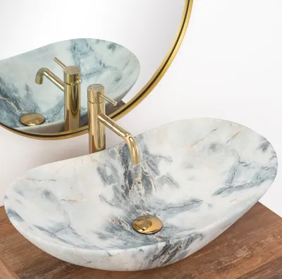 A marble sink with a gold faucet is shown in a bathroom setting, reflected in a round mirror.