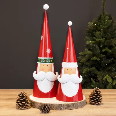 Two Santa Claus decorative figures are displayed on a wooden stand, with pine cones in the foreground and a Christmas tree in the background.