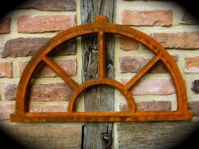 A rusty, arched metal window frame is mounted on a brick wall with wooden beams.