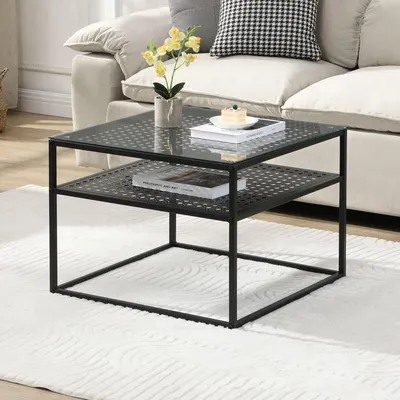 A modern glass and metal coffee table with two shelves, placed in a living room setting next to a sofa.