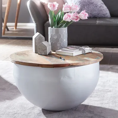 A round coffee table with a wooden top and a white spherical base, decorated with a vase of pink tulips, two small concrete house figurines, and a book.