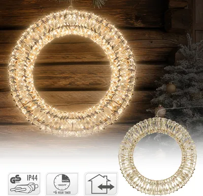 A large illuminated Christmas wreath hangs in front of a wooden wall, with a smaller wreath and a frosted Christmas tree visible in the background. Several icons are displayed at the bottom.