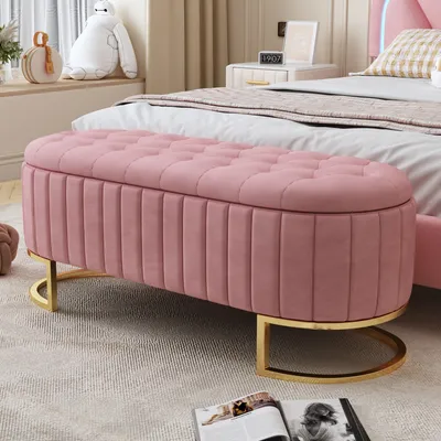 A pink velvet storage bench with gold metal legs is placed in a bedroom setting.