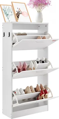 A white shoe cabinet with multiple flip-down drawers, displaying various shoes inside. Two framed pictures and a vase of flowers are on top of the cabinet.