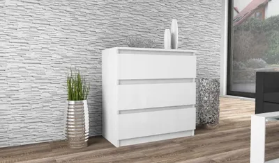 A white, three-drawer dresser is shown in a room with a textured gray wall and a wooden floor. Decorative items, including vases and a plant, are placed around the dresser.