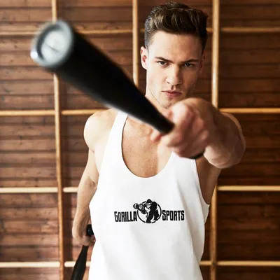 A man in a white tank top with a logo is holding a black baseball bat, looking intensely at the camera. The background is made of wooden planks.