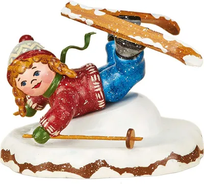 Ein fröhliches Kind mit roten Wangen und Zöpfen trägt einen roten Pullover und blaue Hosen während es auf Skiern eine Bauchlandung im Schnee macht und dabei lächelt