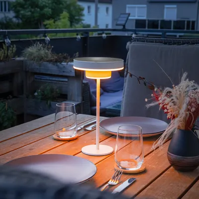 A table lamp is lit on a wooden table on a balcony at dusk.