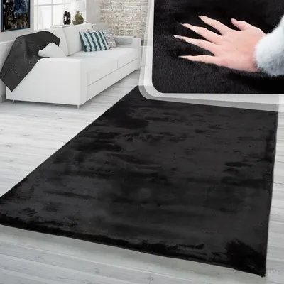 A black shag rug is shown in a living room setting with a white sofa and a hand pressing into the rug to show its softness.