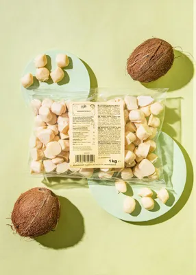 A bag of white chocolate coconut balls with two coconuts and some scattered balls on a light green background.