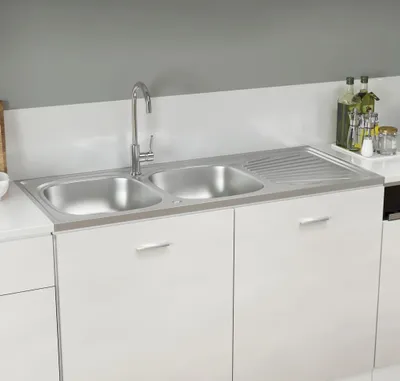A kitchen sink with a double basin and a draining board, set into white cabinets against a gray wall.
