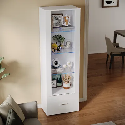 A tall, narrow white display cabinet with glass shelves and a drawer, filled with decorative items and illuminated by blue LED lights. It is placed in a room next to a sofa and a doorway.