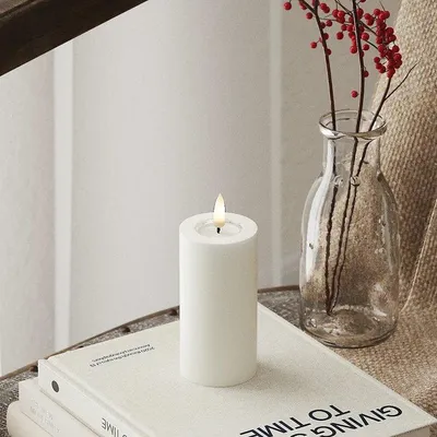 A white LED candle with a lit flame sits on top of a stack of books, next to a glass vase with red berries.