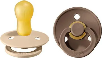 Two BIBS pacifiers are shown against a white background. One is beige with a yellow nipple, and the other is brown with a gold ring and a brown nipple.
