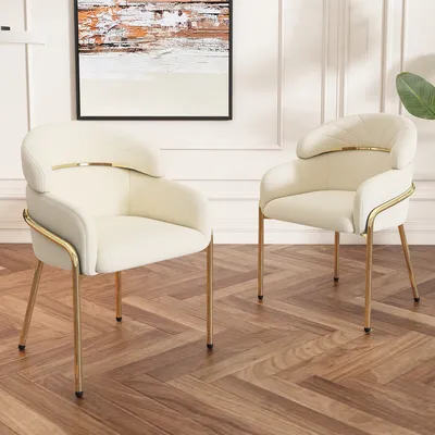 Two beige armchairs with gold legs are placed on a wooden floor in a room with white paneled walls and a painting on the wall.