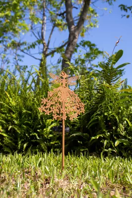 A metal fairy garden stake is shown in a garden setting with green plants and a blue sky.