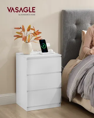 A white nightstand with a vase of flowers and a phone charging on top, next to a bed with a person sitting up.