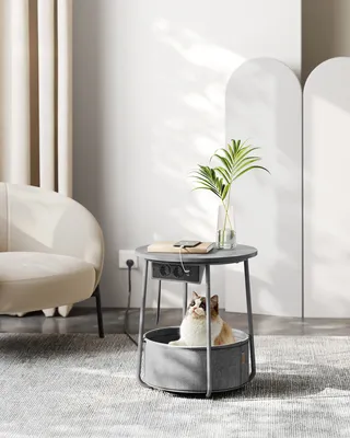 A cat is sitting in a pet bed integrated into a round side table with built-in power outlets and USB ports. The table has a plant in a vase on top, and a phone is charging. A chair is visible in the background.