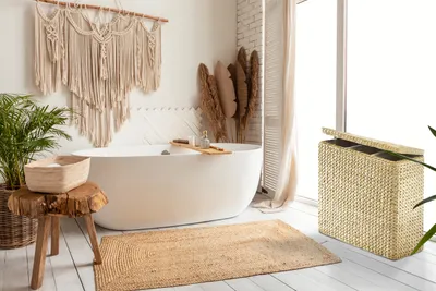 A bright and airy bathroom with a freestanding bathtub, macrame wall hanging, plants, and a woven laundry sorter.