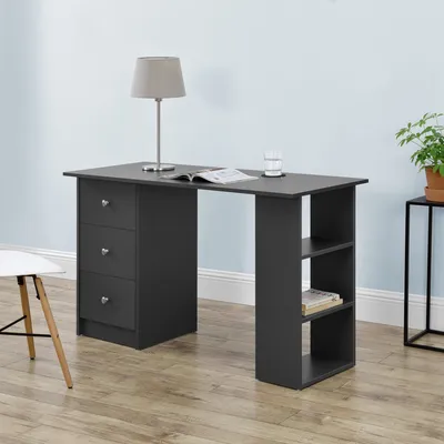 A dark gray desk with drawers and shelves, a lamp, and a glass of water on top, set against a light blue wall and wooden floor.