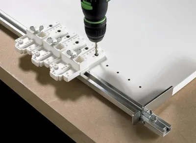 A drill is being used with a jig to drill holes in a white board, which is mounted on a metal rail.