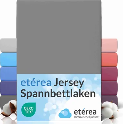 A folded grey jersey fitted sheet is shown, with stacks of other colored sheets visible behind it. Cotton bolls are in the foreground.