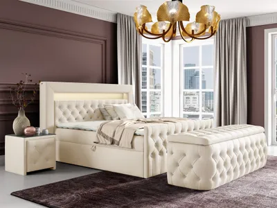 A luxurious bedroom setting with a large boxspring bed, a matching ottoman, and a bedside table, illuminated by a chandelier and ambient lighting.