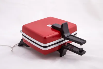 A red waffle iron with black accents and feet, shown on a white background.