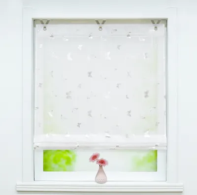 A white Roman blind with a butterfly pattern is shown hanging in a window. Two pink flowers in a small vase are on the windowsill below.
