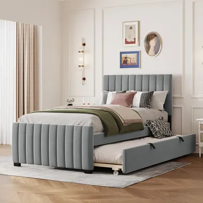 A grey upholstered bed with a pull-out trundle bed underneath, set in a bedroom with decorative wall art and curtains.