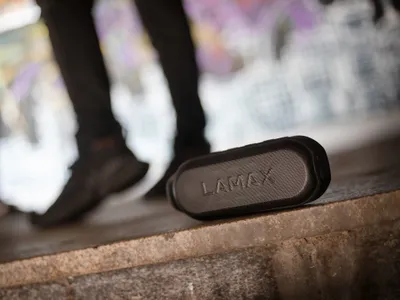 A black portable speaker sits on a concrete ledge with a person's legs in black pants and shoes blurred in the background.