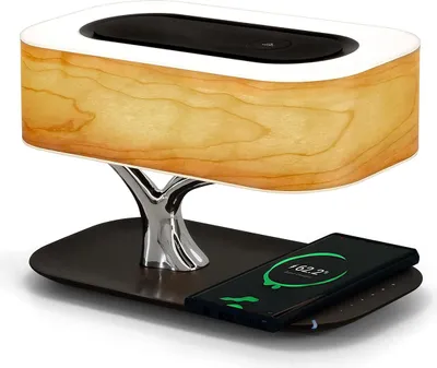 A modern bedside lamp with a wood-grain finish, a chrome tree-like stand, and a black base, with a smartphone charging on the base.