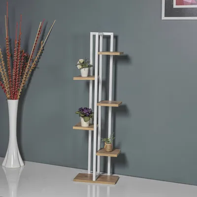 A tall, narrow plant stand with multiple shelves is shown against a gray wall. A white vase with dried branches is to the left.