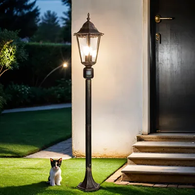 A rustic outdoor lamp stands in a garden at dusk, with a small cat sitting on the grass in front of it.