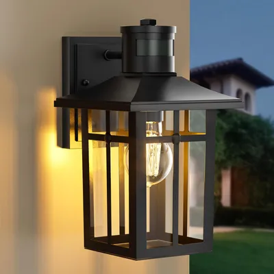 A black outdoor wall lamp with a motion sensor is mounted on a beige wall. The lamp has a vintage design with glass panels and an Edison-style bulb. The background shows a blurred view of a house and greenery at dusk.