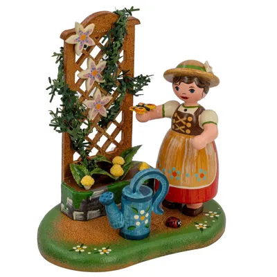 Ein kleines Mädchen in traditioneller Kleidung steht neben einer blühenden Rankhilfe und hält eine Gießkanne sowie eine Blume in der Hand während eine Marienkäferfigur am Boden sitzt