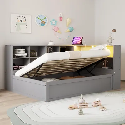 A grey bed with its storage compartment open, revealing the mattress and storage space. The bed is in a child's room with decorations on the wall and toys on the floor.
