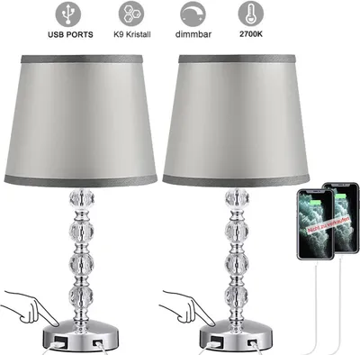 Two identical crystal table lamps with gray fabric shades are shown, with icons and text above indicating features like USB ports, dimmability, and color temperature. Phones are shown charging from the lamps.