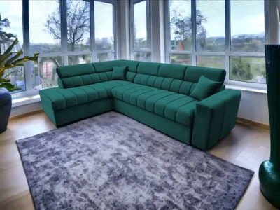 A green sectional sofa is placed on a gray rug in a room with large windows overlooking a landscape.