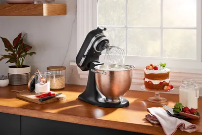 A black KitchenAid stand mixer with a copper bowl is shown on a wooden countertop next to a small cake and a jar of milk.