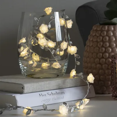 A string of LED lights shaped like roses is draped inside a glass vase, which sits on top of two books. A textured vase is visible in the background.