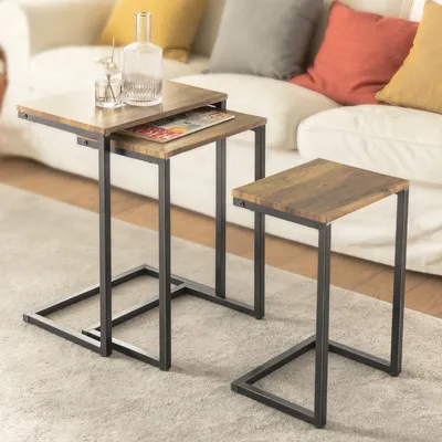 Two nesting side tables with wood-look tops and dark metal frames are placed on a rug in front of a sofa with colorful cushions.