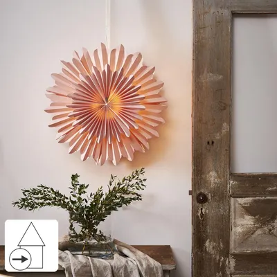 A pink paper star lantern hangs on a wall next to a rustic wooden door. Below the lantern, a vase of greenery sits on a wooden bench.