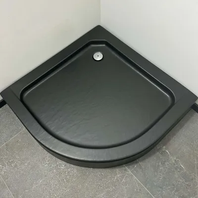 A black corner shower tray with a drain in the center, placed in a bathroom setting with gray tiled flooring and light gray walls.