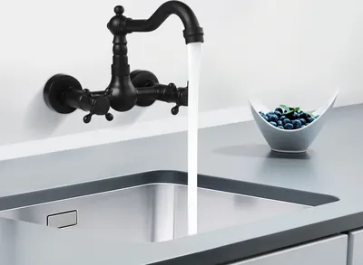 A black wall-mounted faucet is running water into a stainless steel sink. A small bowl of blueberries is on the countertop next to the sink.