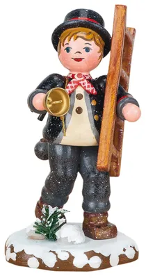 Ein fröhlicher Schornsteinfeger in traditioneller Kleidung hält eine Leiter und eine goldene Glocke während er auf einem schneebedeckten Sockel steht und ein kleines grünes Tannenbäumchen neben ihm wächst