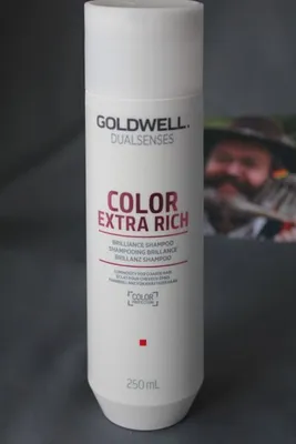 A white bottle of Goldwell Dualsenses Color Extra Rich Brilliance Shampoo is shown against a gray background. A blurred image of a man with a beard wearing a hat is visible in the background.