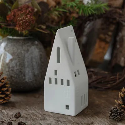 A white ceramic house-shaped lantern sits on a wooden surface, surrounded by natural elements like pinecones and dried foliage.