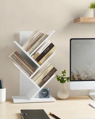 A white tree-shaped bookshelf filled with books is placed on a desk next to a computer monitor and a small plant.